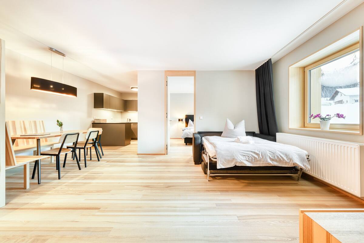 A modern hotel room with a wooden floor, a bed with a checkered duvet, a dining area with a table and chairs, and a view of a snowy landscape through a window.