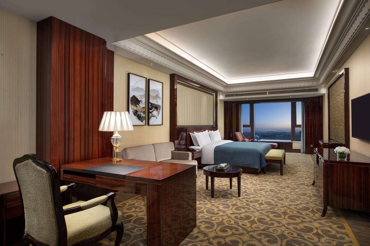 A hotel room with a desk, two seating areas, a bed, and a large window with a view.