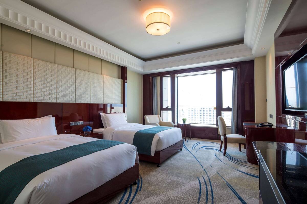 A hotel room with two beds, a television, a desk, and a large window.