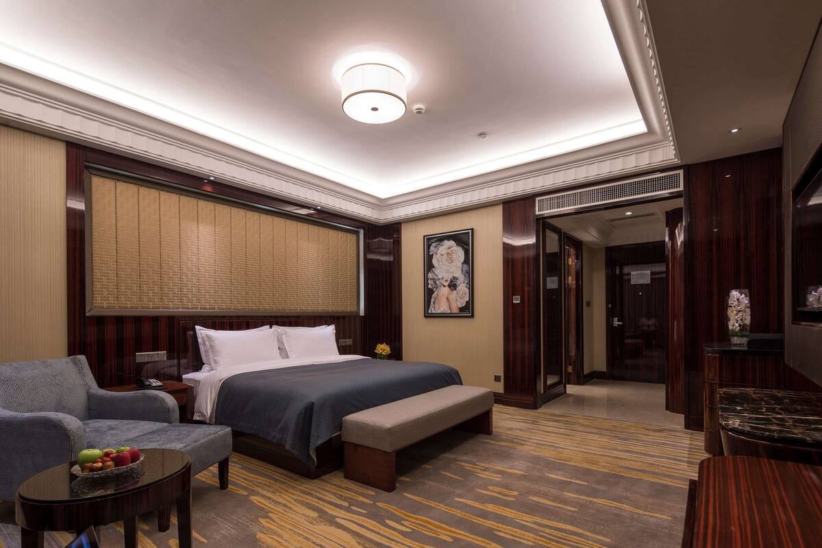 A hotel room with a bed, a sofa, a small table with fruit, a bench, and a desk with a vase of flowers.
