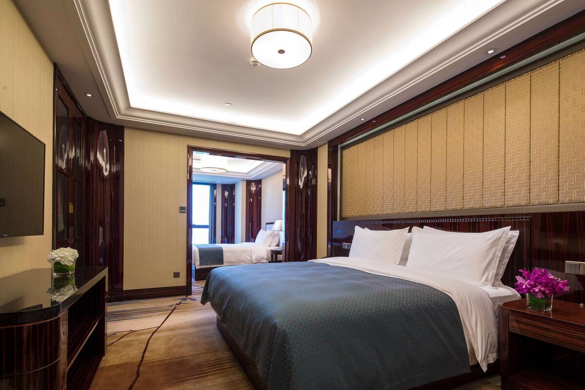 A hotel room with a large bed, a flat-screen TV, a desk with a vase of flowers, and a view of the hallway.