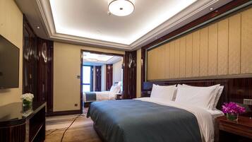 Habitación de hotel con una cama grande, televisor de pantalla plana, escritorio con un jarrón de flores y vistas al pasillo.