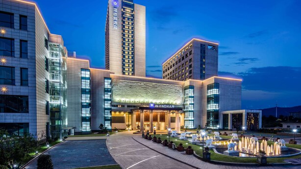 A modern hotel with a prominent entrance, a central fountain, and a tall building with illuminated windows. - Wyndham Zhuji West (Shaoxing)