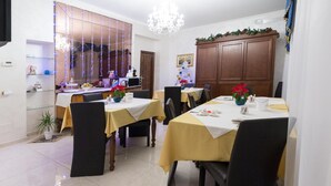 A dining area with tables set for four, a kitchen in the background, and a chandelier. - Roma Royal Residence (Rome)