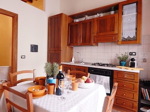 Une cuisine avec des meubles en bois, une cuisinière à gaz, un évier et une table à manger, mise avec des bols, des verres à vin et un panier de fruits.