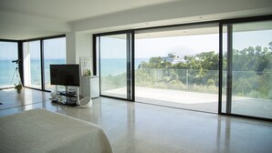 A spacious room with a large sliding glass door leading to a balcony with a view of the sea and greenery. - The Cliff House (Carthage)