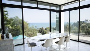 A modern dining area with a large oval table and six chairs, large floor-to-ceiling windows with a view of the sea and greenery. - The Cliff House (Carthage)