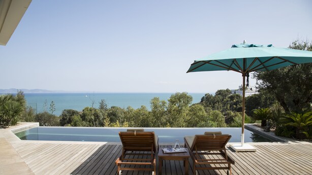 A wooden deck with two lounge chairs and a table, overlooking a pool and a scenic view of the sea and hills. - The Cliff House (Carthage)