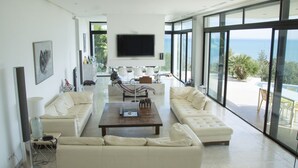 A spacious living room with a large TV, white sofas, a coffee table, and a view of the ocean through large windows. - The Cliff House (Carthage)