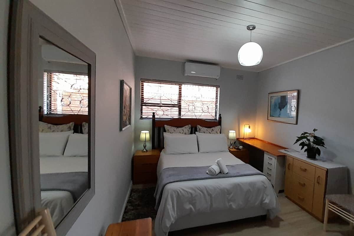 A bedroom with a bed, bedside tables, a desk, a chair, a mirror, and a window with blinds.