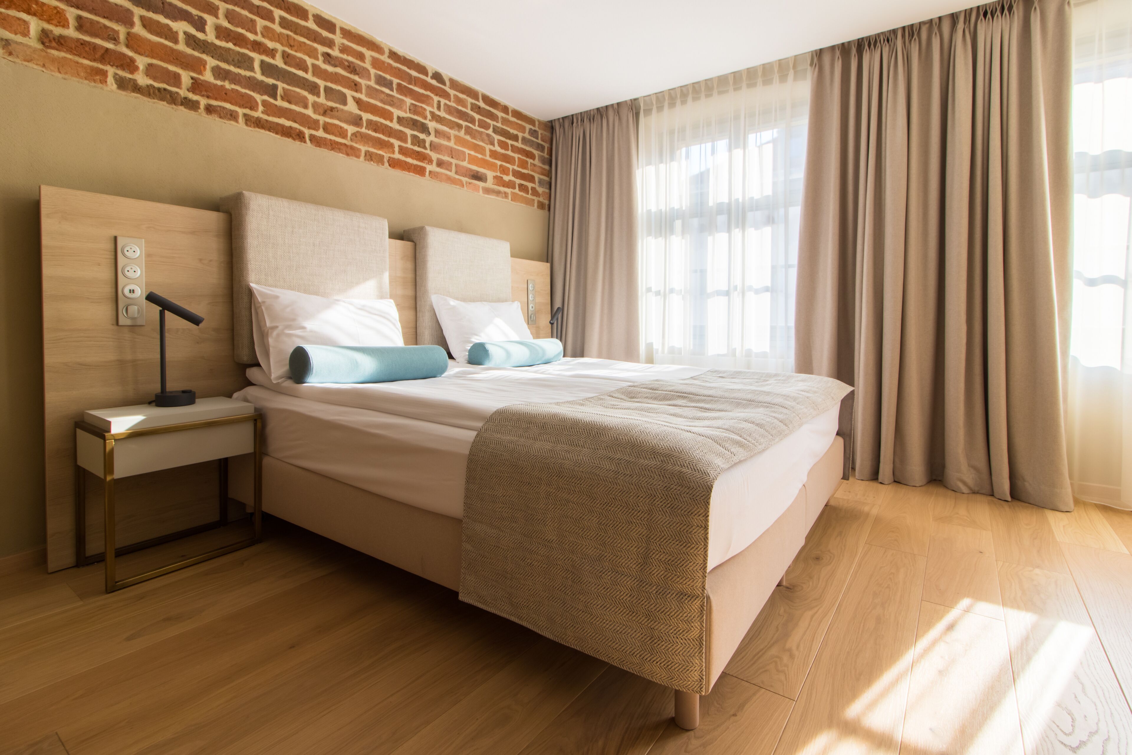A modern hotel room with a brick wall, a wooden floor, a bed with beige bedding, a nightstand with a lamp, and a window with curtains.