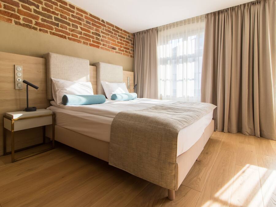 Ett modernt hotellrum med en tegelvägg, ett trägolv, en säng med beige sängkläder, ett nattduksbord med en lampa och ett fönster med gardiner.