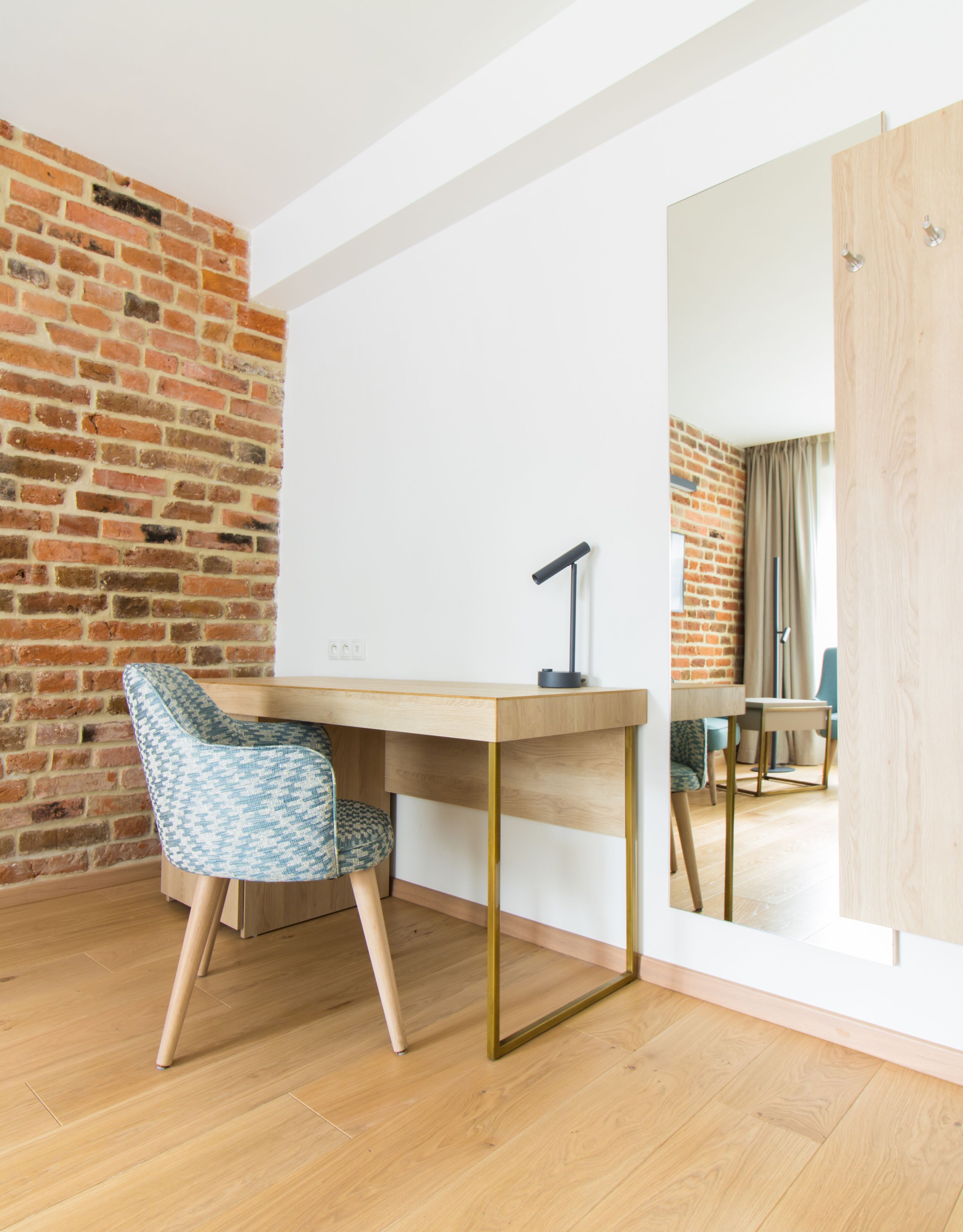 A modern room with a brick wall, a wooden desk, a patterned chair, and a large mirror reflecting another room.