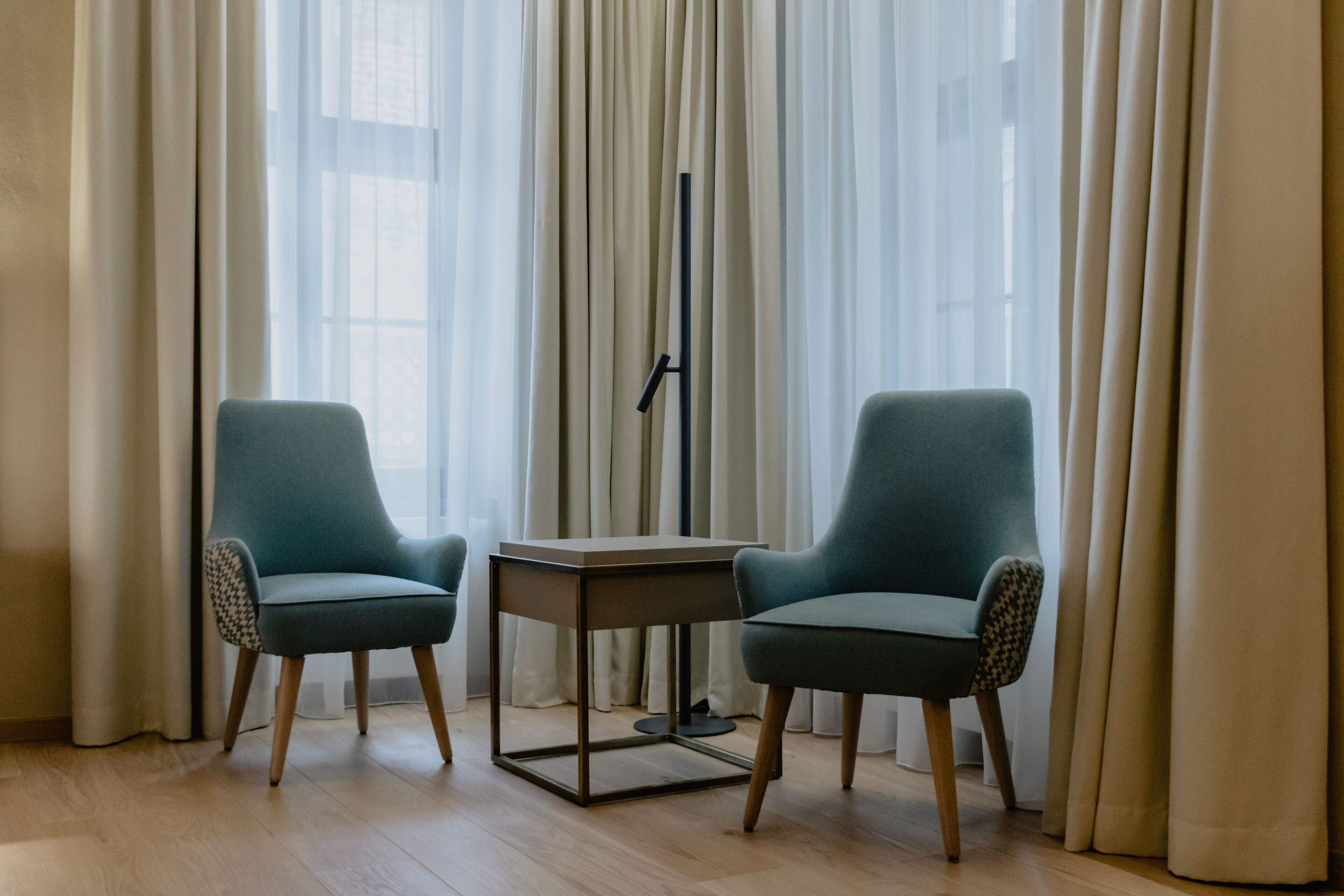 A room with two teal armchairs, a small wooden table, and sheer curtains.