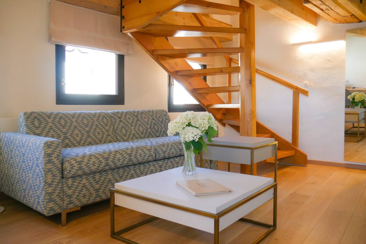 Un salón con un sofá estampado, una mesita blanca con un florero y una escalera de madera.