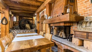 Un interior rústico de madera con pared de piedra, vigas de madera, chimenea y una mesa con sillas.