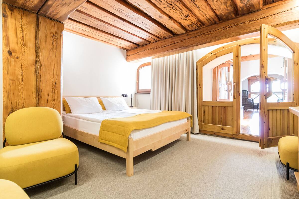 Un dormitorio con techo de madera, una cama con ropa de cama blanca, un sillón amarillo y una puerta de madera que da a un espacio exterior.