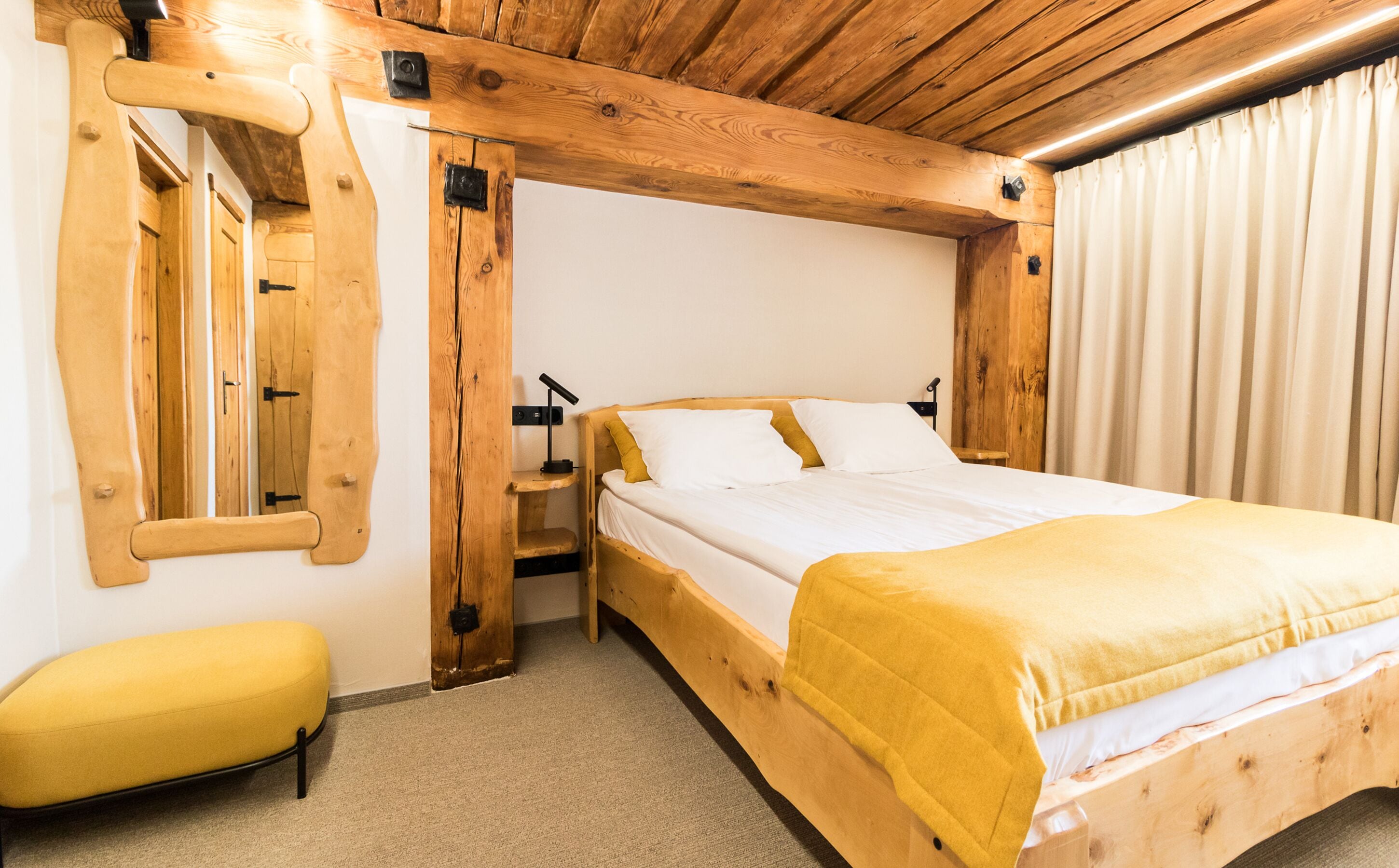 Un dormitorio con estructura de madera, una cama, un banco, un espejo y un ventanal con cortinas.