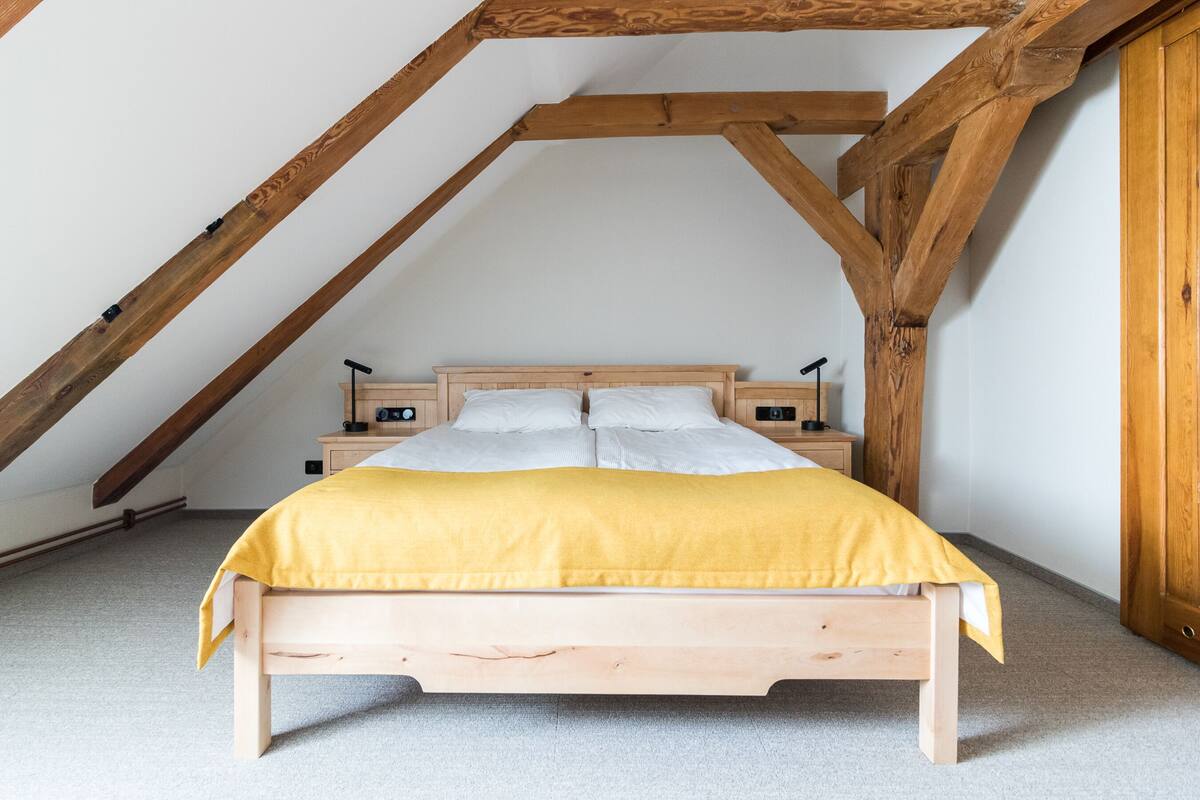 Un dormitorio con estructura de cama de madera, colcha amarilla, dos mesitas de noche con lámparas y un techo inclinado con vigas de madera a la vista.