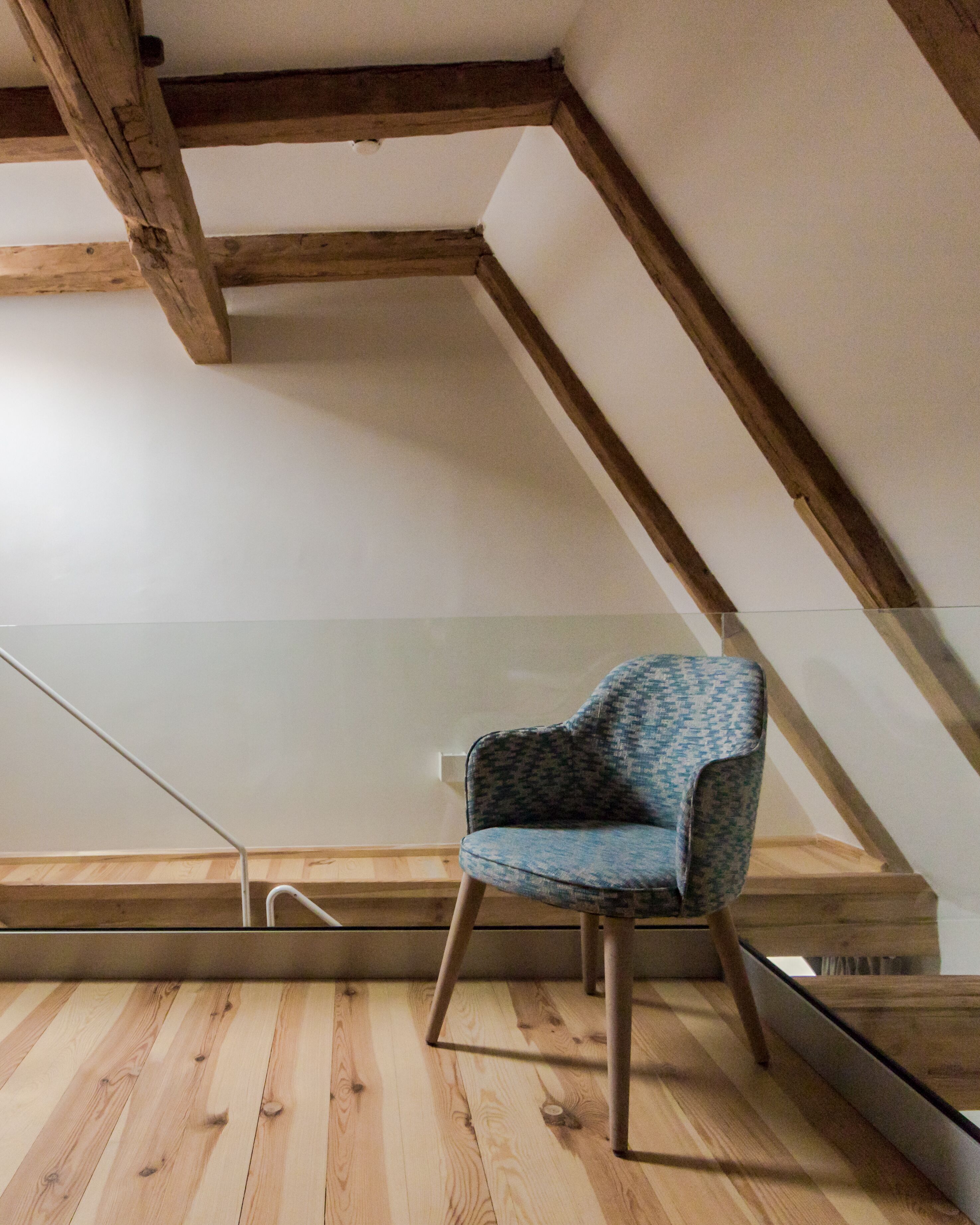 A room with a wooden floor, a blue patterned chair, and exposed wooden beams on the ceiling.