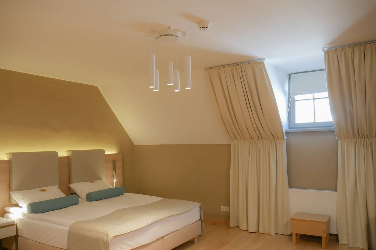Un dormitorio moderno con una cama grande, mesitas de noche, un taburete de madera y una ventana con cortinas.