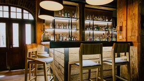 Una zona de bar con sillas de madera y una barra, estantes con botellas, y un amplio ventanal con luz natural.