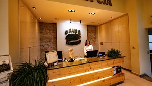 Un comptoir d’accueil avec une enseigne indiquant « RECEPCIA » et « OSADA » au-dessus. Une femme se tient derrière le comptoir, à côté duquel se trouvent un ordinateur et une plante.
