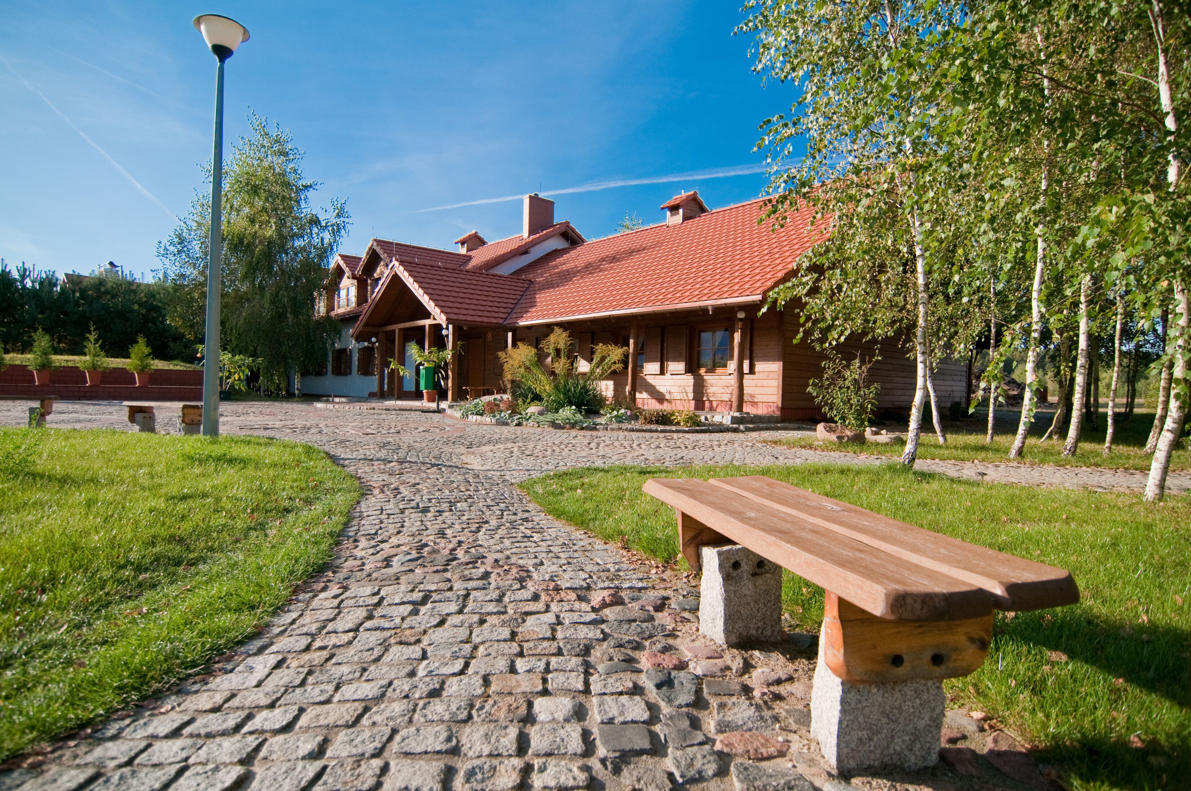 Ein Kopfsteinpflasterweg führt zu einem Holzhaus mit rotem Ziegeldach, umgeben von Grünpflanzen und einer Bank.