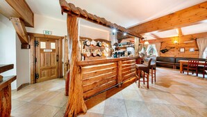 Un coin bar rustique, avec un comptoir en bois, un plafond en bois et un sol carrelé. On y trouve une porte en bois avec une enseigne, un banc en bois et une table avec des chaises.