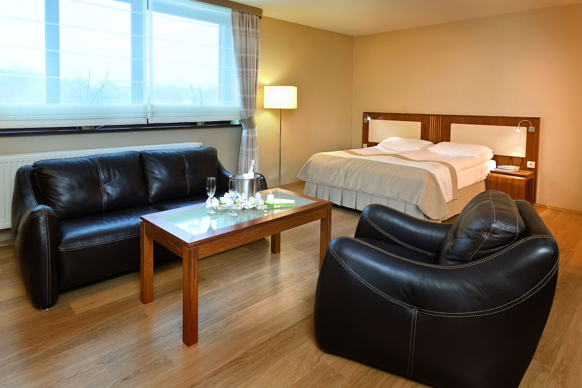 Una habitación de hotel con un sofá de cuero, una mesa de centro de madera, una cama con edredón blanco y una mesita de noche con lámpara.