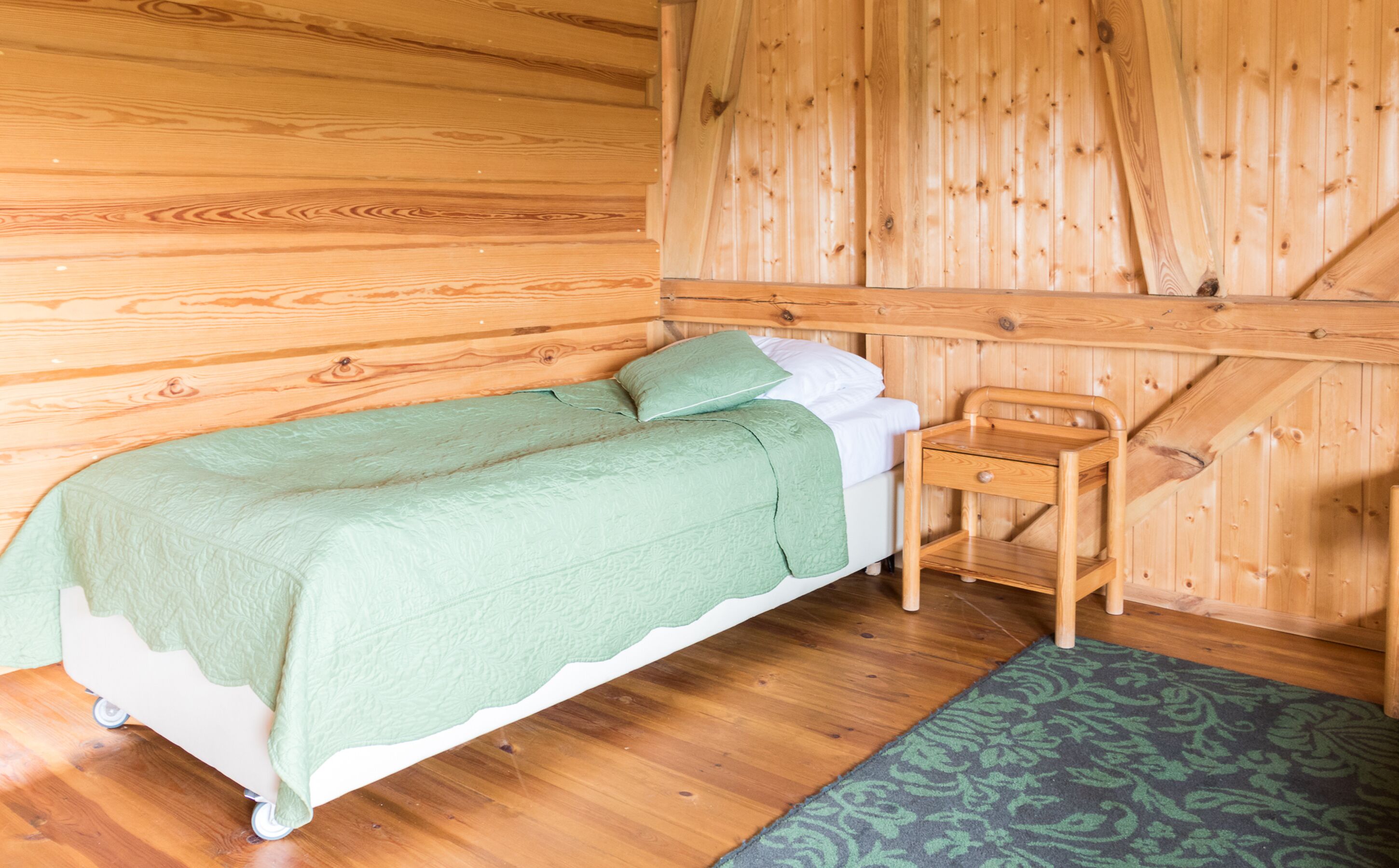A wooden cabin room with a bed, a small wooden chair, and a patterned rug.