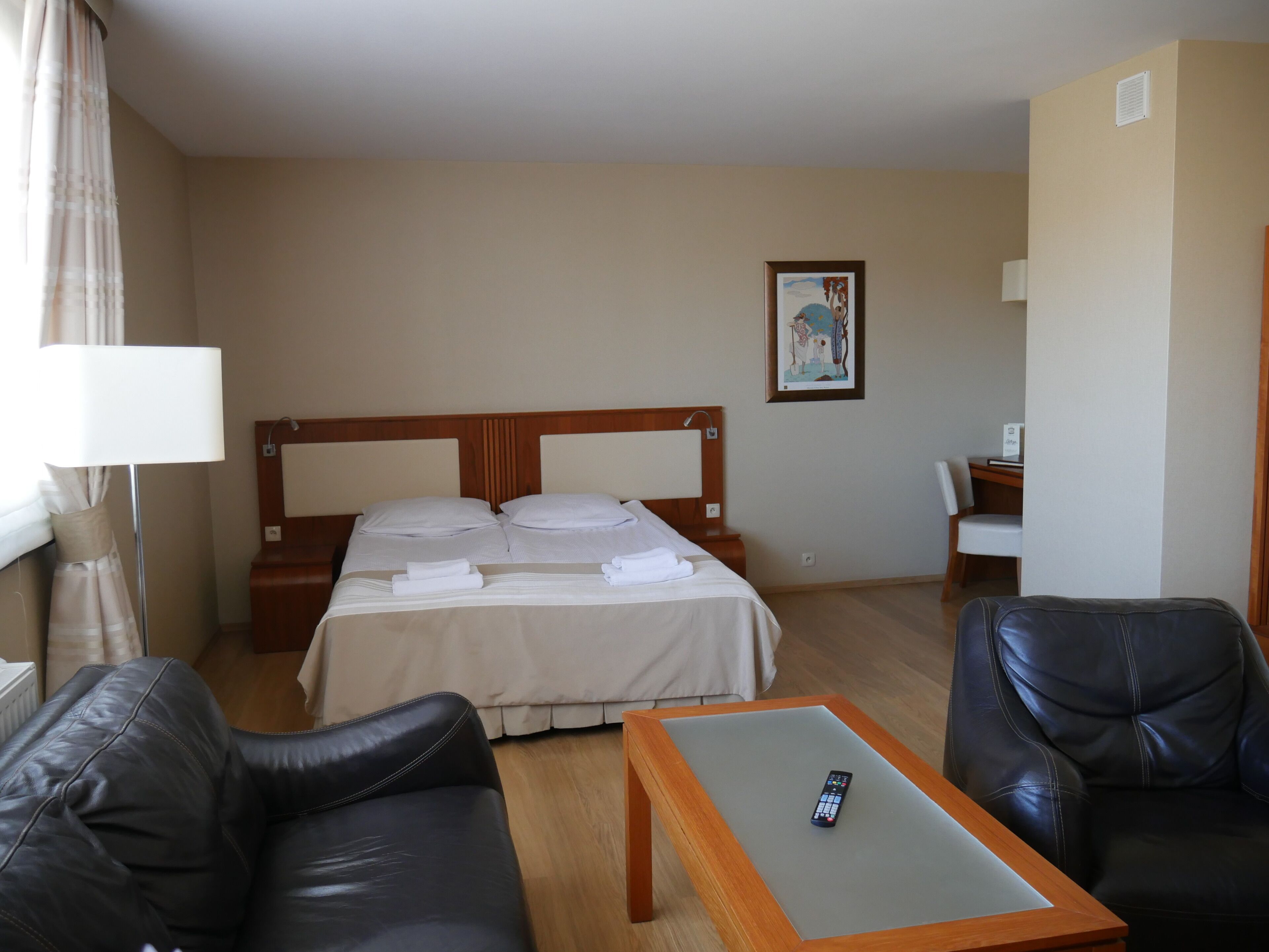 A hotel room with a bed, a desk, a chair, a remote control on the desk, and a painting on the wall.