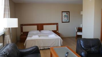 A hotel room with a bed, a desk, a chair, a remote control on the desk, and a painting on the wall.