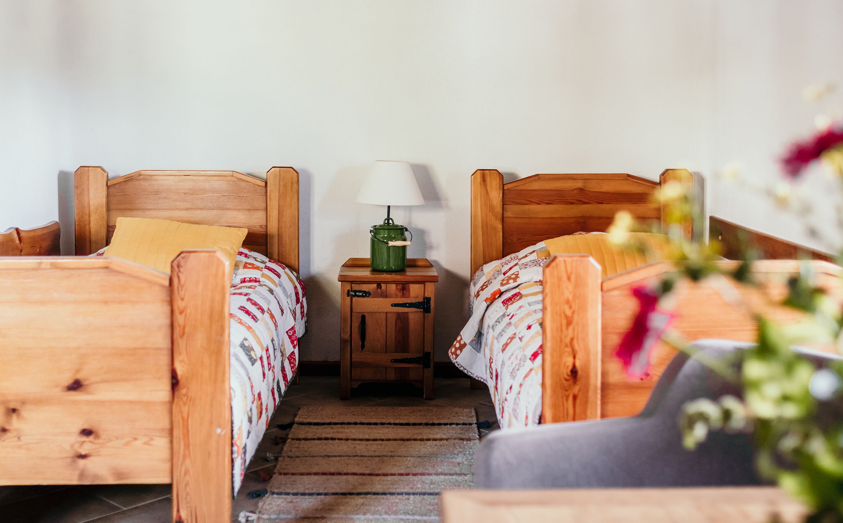 Una habitación pequeña y acogedora con dos camas de madera, cada una con un cojín y una colcha estampada. Un pequeño mesita de noche de madera con una lámpara verde y una blanca se encuentra entre las camas.