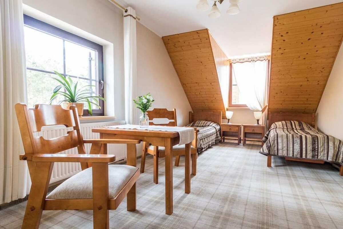 A cozy room with a sloped ceiling, a bed with a striped bedspread, a wooden table with chairs, and a window with curtains.