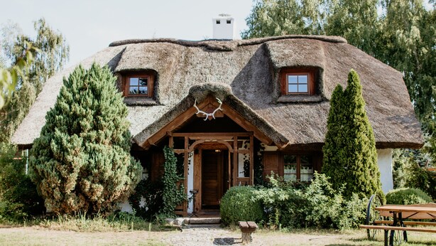 Un petit chalet au toit de chaume, doté d’une porte d’entrée en bois et de deux fenêtres, entouré de verdure.