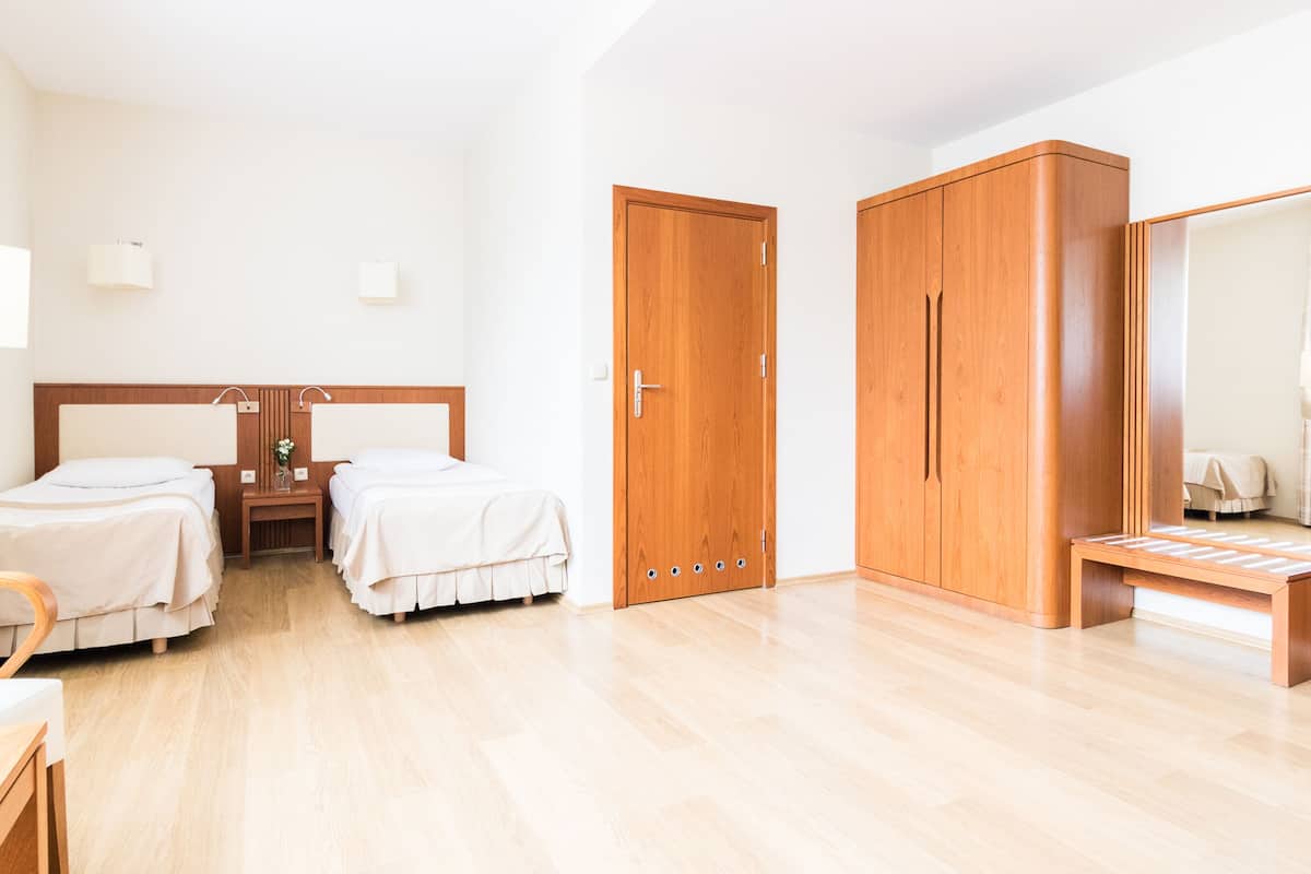 Habitación de hotel con dos camas individuales, un armario de madera, una mesita de noche con lámpara y un escritorio pequeño con silla.