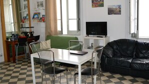 A living room with a checkered floor, a green sofa, a black leather couch, a dining table with chairs, a TV on a stand, and a window with curtains. - Hostel Via Zara (Palermo)