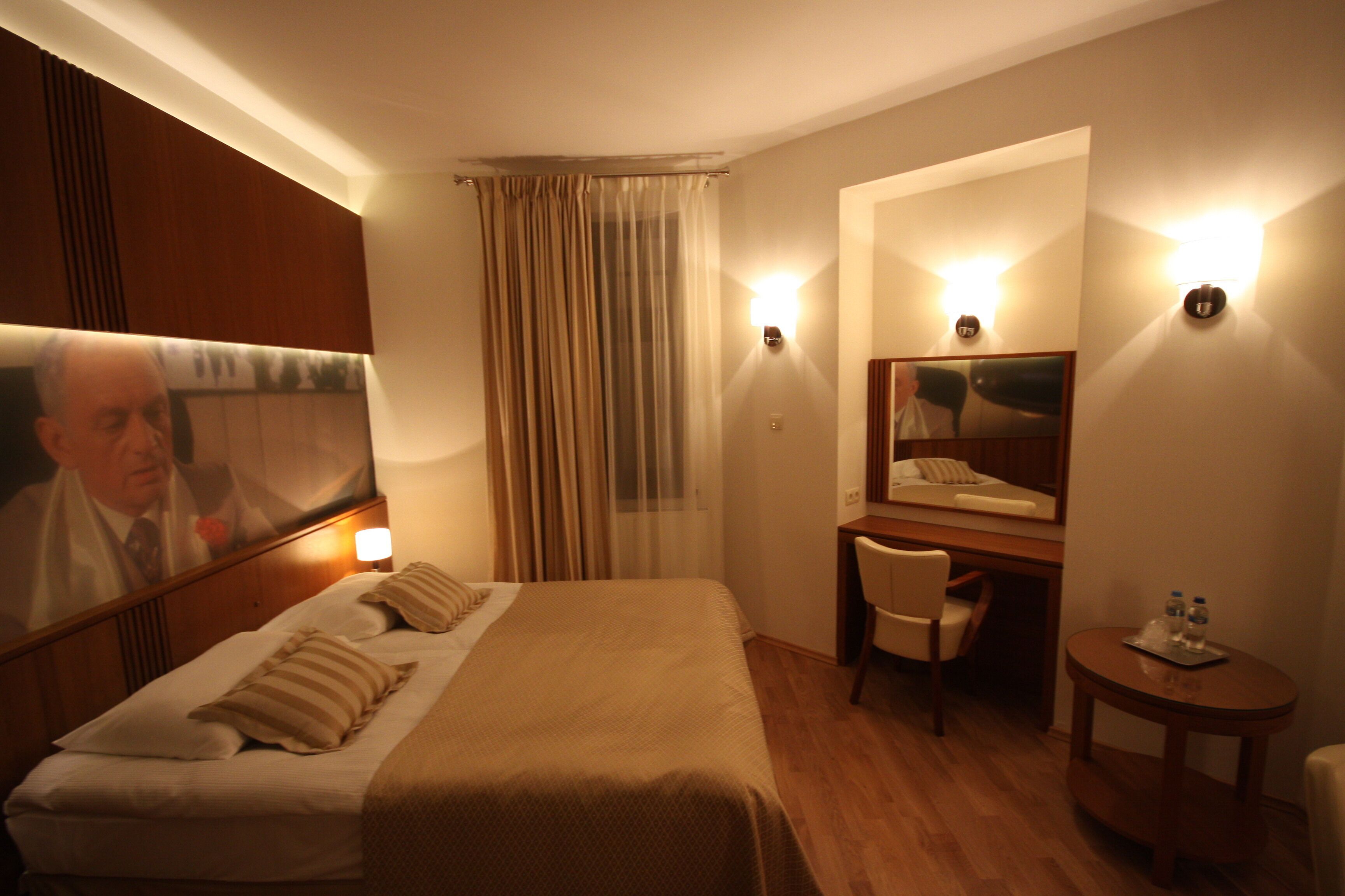 Ein Hotelzimmer mit Bett, Schreibtisch, Stuhl, einem kleinen Tisch mit Wasserflaschen, einem Spiegel und einem Vorhang.