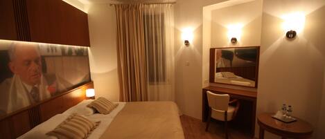 Ein Hotelzimmer mit Bett, Schreibtisch, Stuhl, einem kleinen Tisch mit Wasserflaschen, einem Spiegel und einem Vorhang.