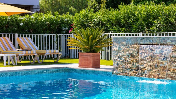 A swimming pool with a tiled edge, surrounded by a white fence and a row of striped lounge chairs.