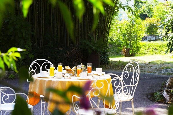An outdoor dining area with a round table set for breakfast, surrounded by white metal chairs, in a lush garden setting. - Hotel Du Bosquet (Pegomas)