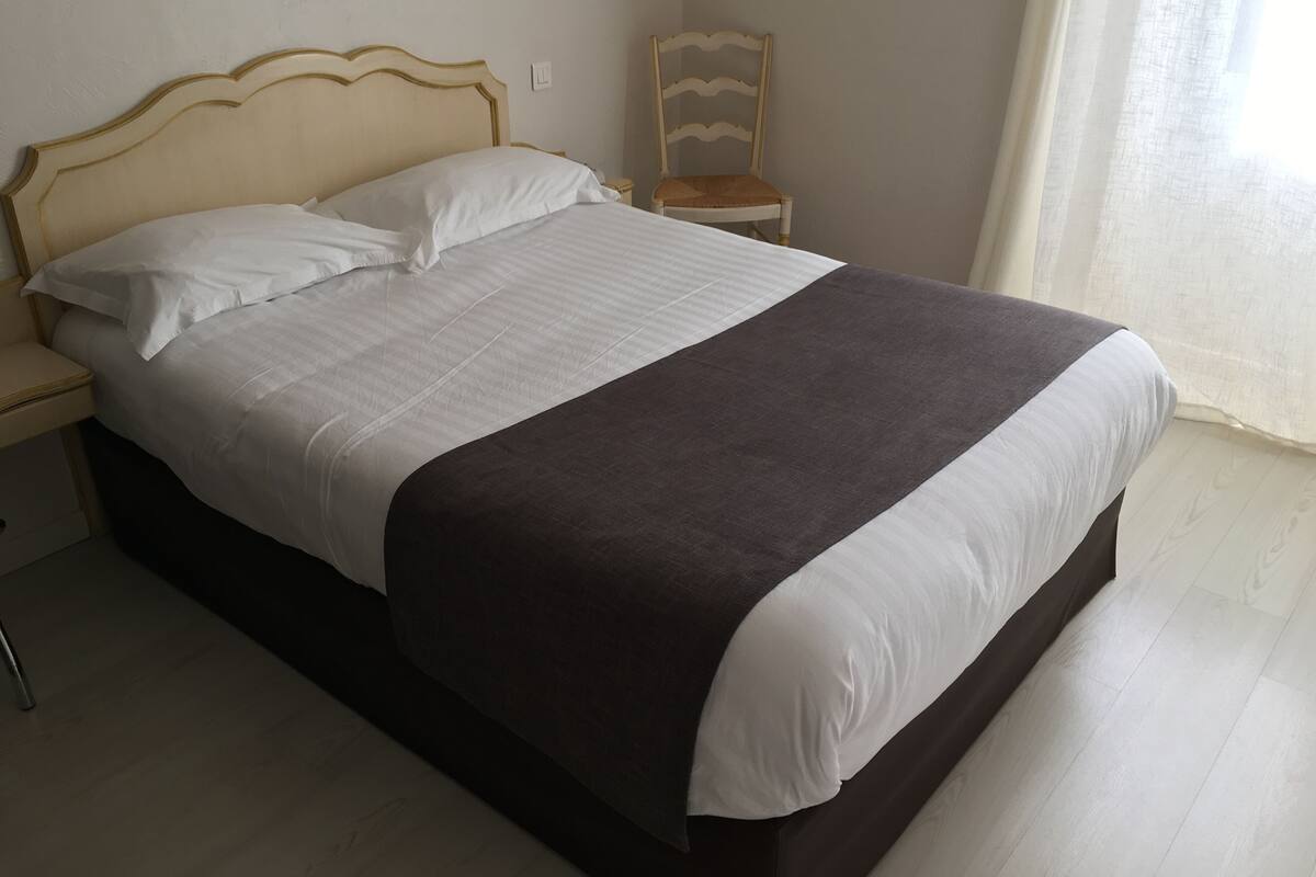 A neatly made bed with white and dark bedding, a wooden headboard, and a small bedside table with a chair.