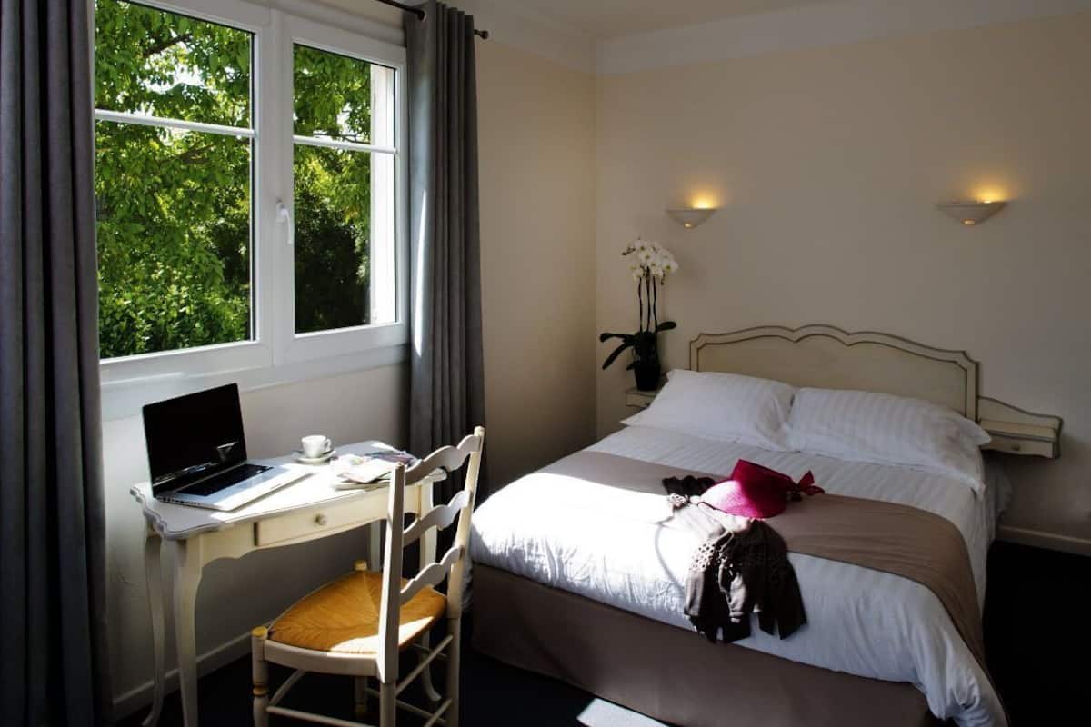 A hotel room with a bed, a desk with a laptop, a chair, and a window with curtains.