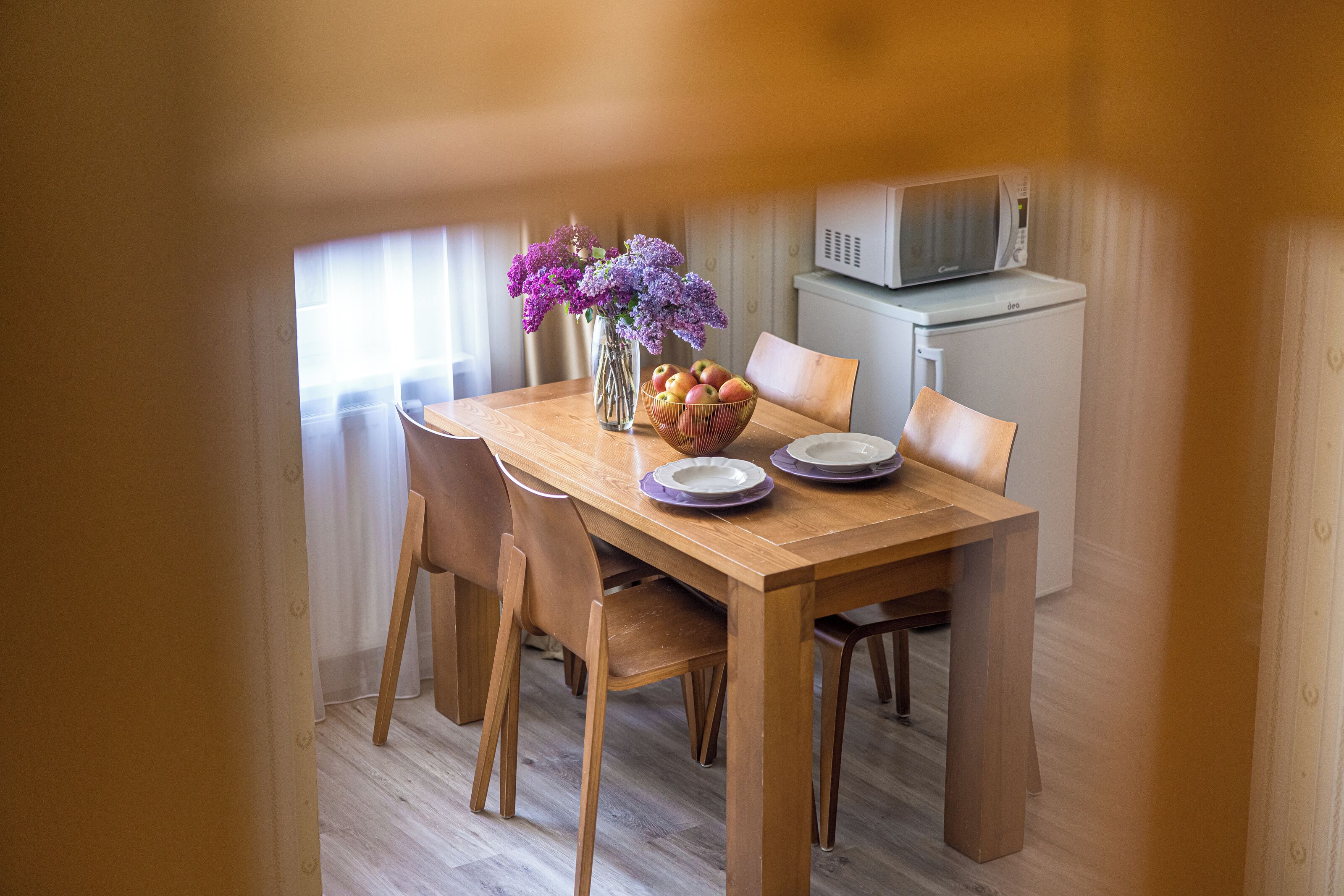 A dining area with a wooden table, chairs, a vase with purple flowers, and a bowl of fruit.