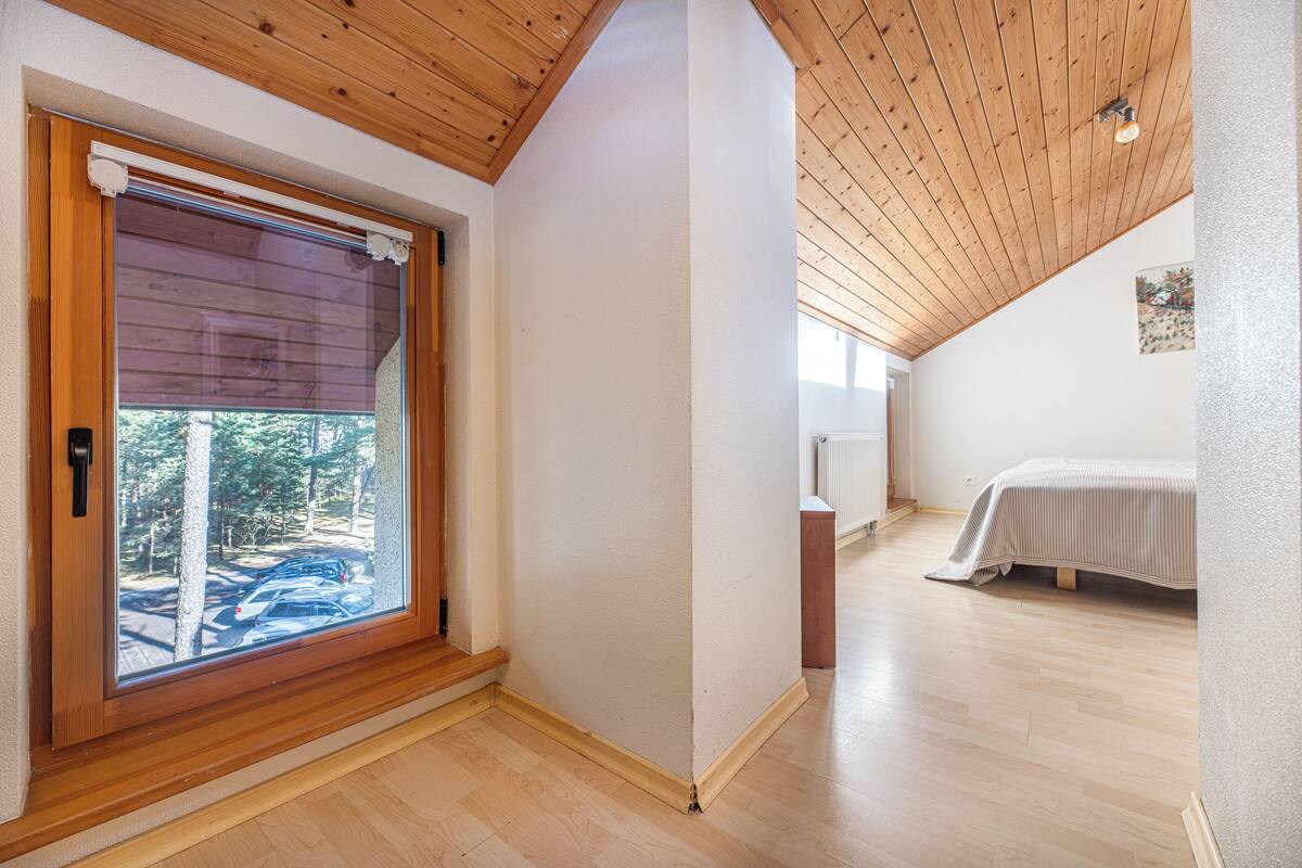 A room with a wooden ceiling, a large window with blinds, a bed with a white bedspread, and a radiator.