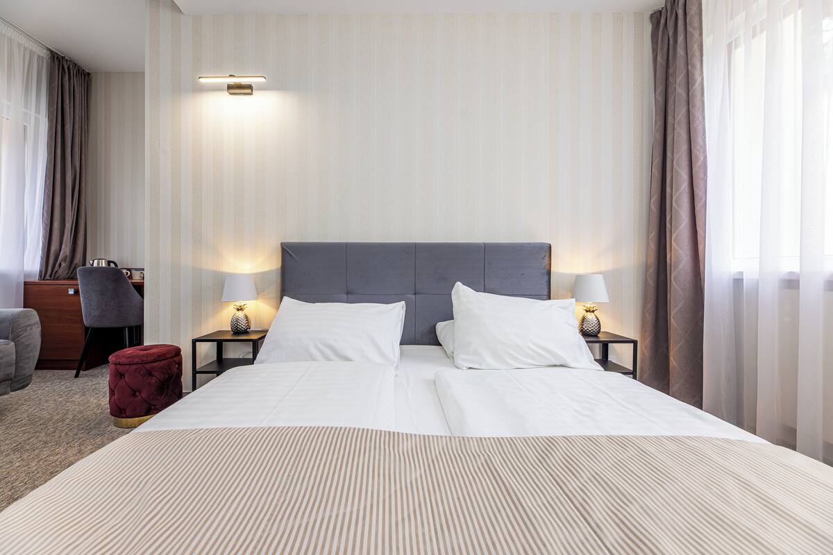 A hotel room with a large bed, two bedside tables with lamps, a red ottoman, a desk with a chair, and a window with curtains.