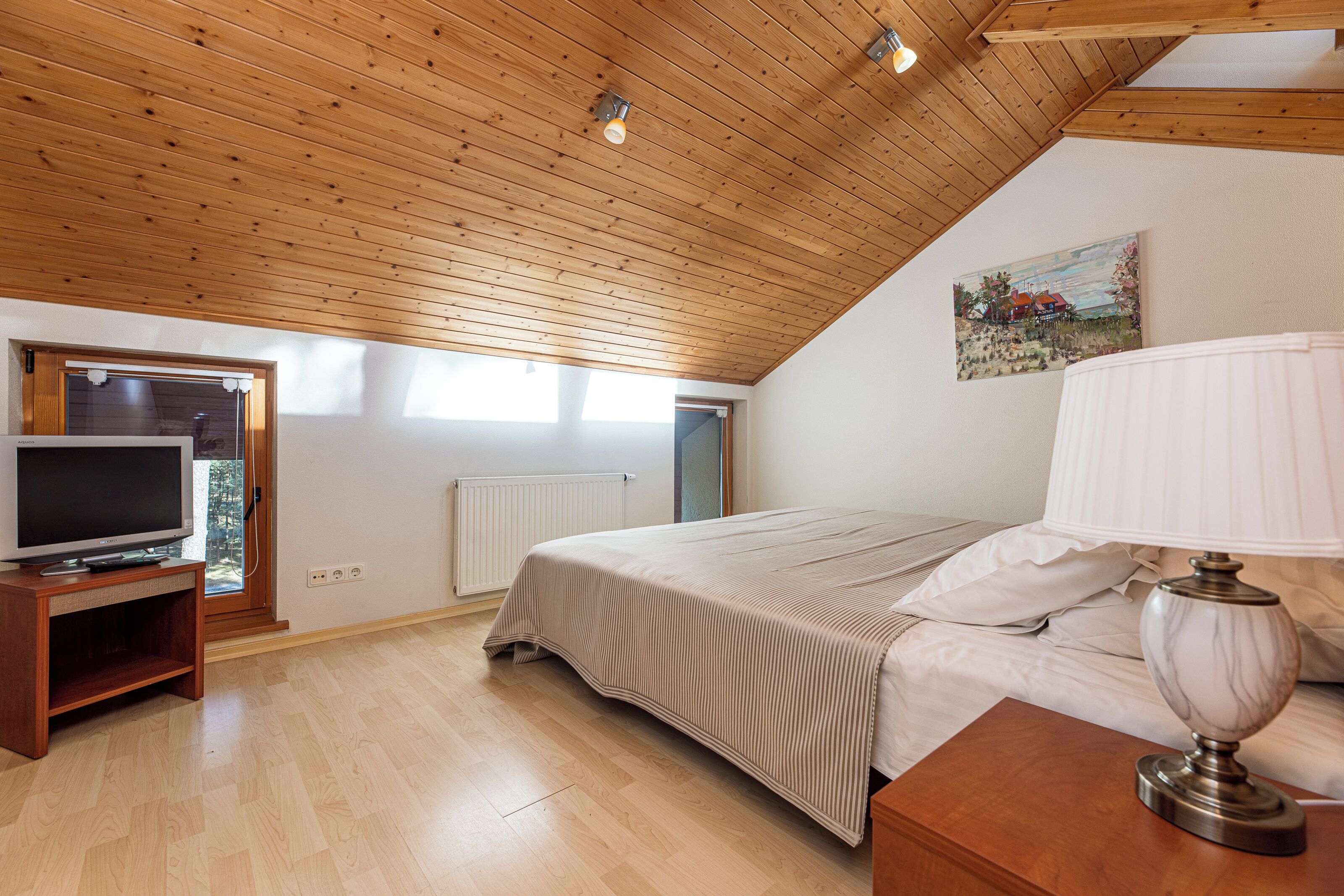 Une chambre avec un plafond en bois, un lit, un téléviseur posé sur un meuble et un tableau accroché au mur.