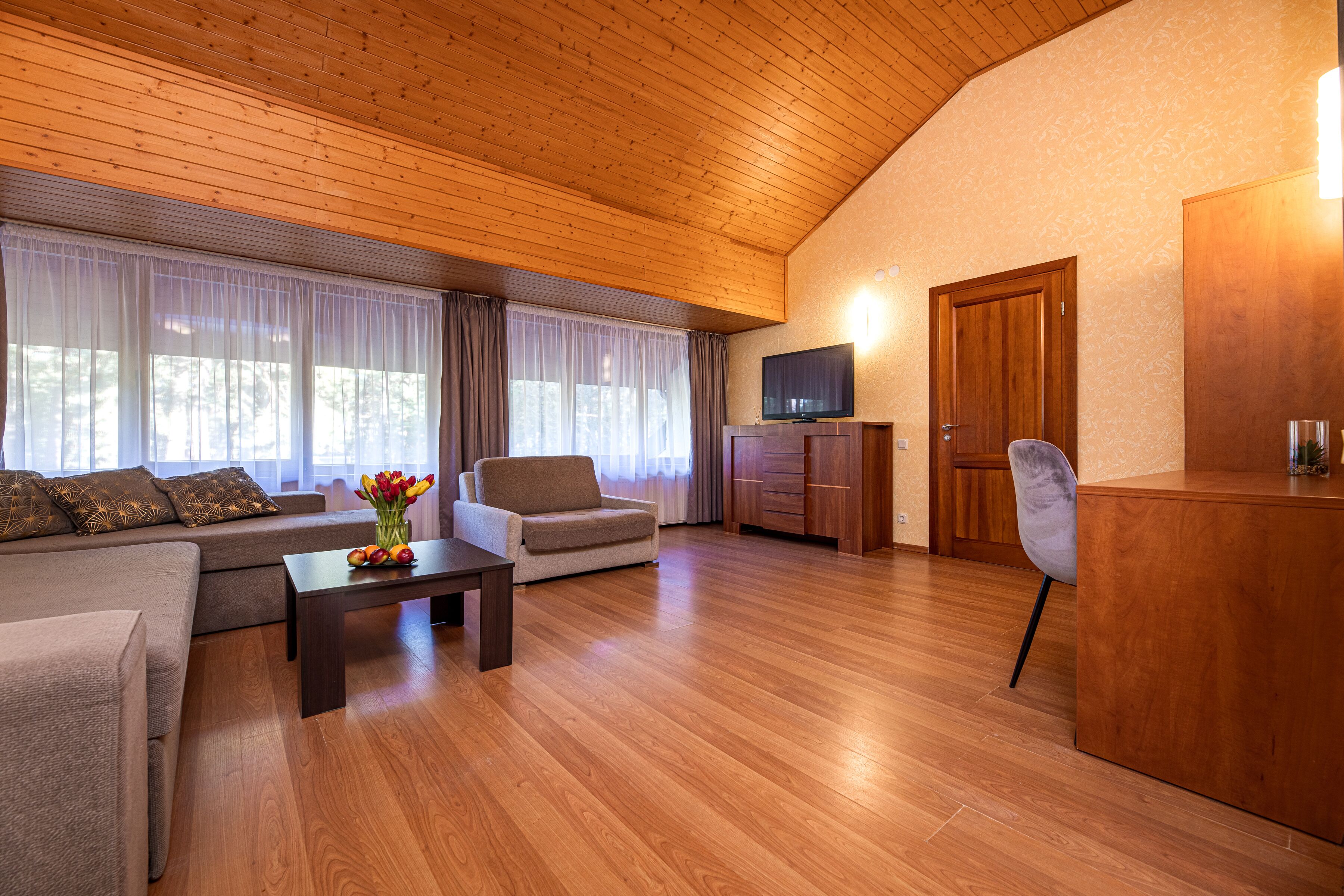 A modern living room with a wooden ceiling, a sofa set, a coffee table with flowers, a TV, and a wooden cabinet.