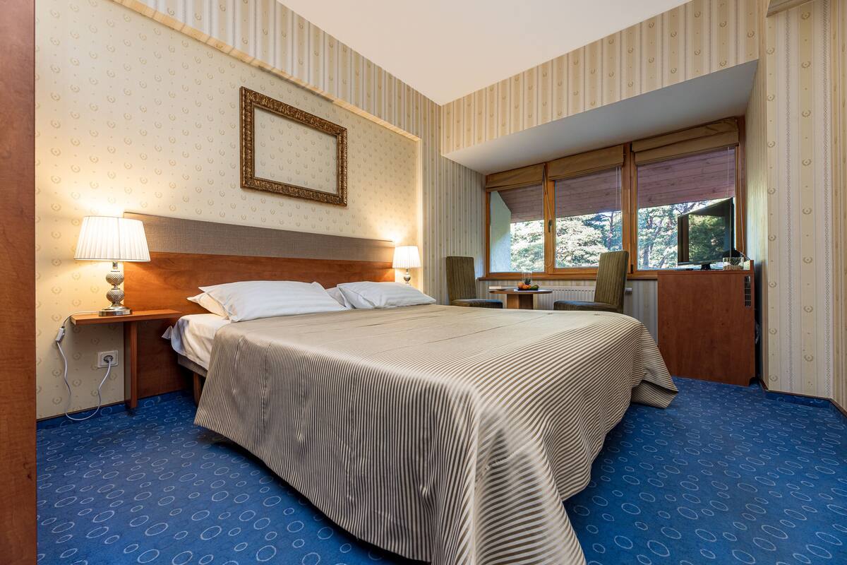 A hotel room with a large bed, two bedside lamps, a wooden headboard, a window with blinds, and a desk with a chair.